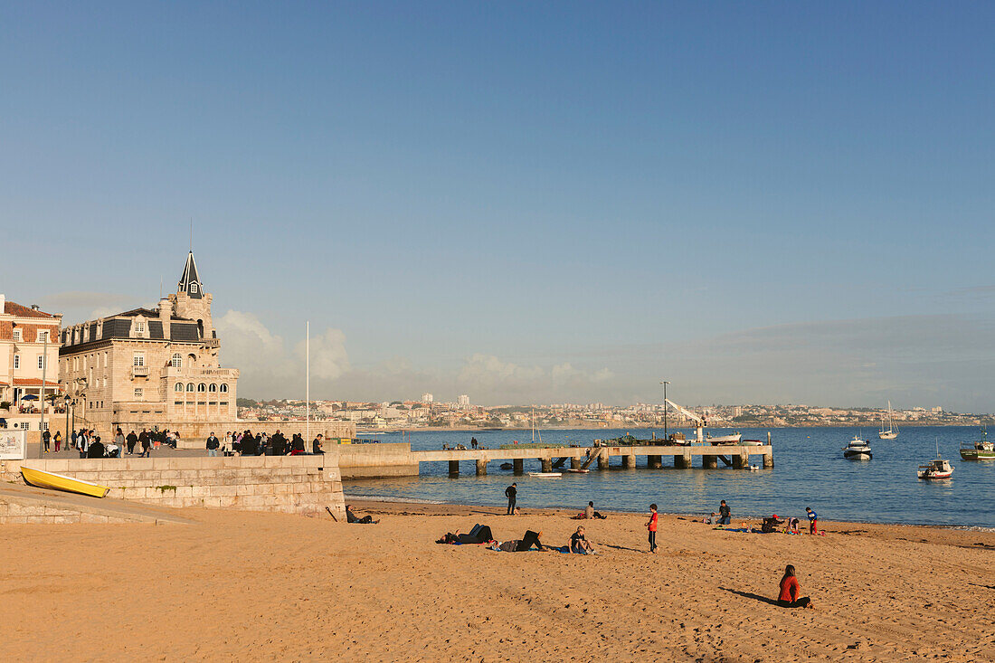 Menschen genießen die Uferpromenade von Cascais, Lisboa, Portugal, mit einem Strand und Booten im Hafen, Lissabon, Portugal