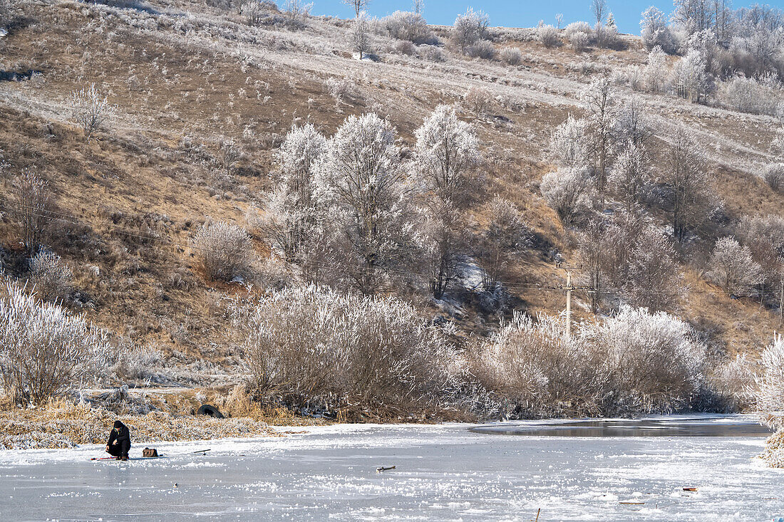 Mann fischt durch ein Loch im Eis eines zugefrorenen Flusses im ländlichen Siebenbürgen,Rumänien,Siebenbürgen,Rumänien