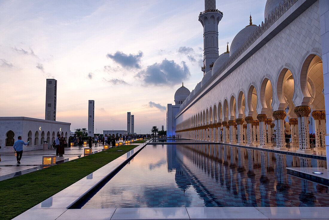 Besucher gehen an einem der spiegelnden Becken der Großen Moschee in Abu Dhabi, VAE, vorbei und fotografieren es, während der Sonnenuntergang in die Nacht übergeht, Abu Dhabi, Vereinigte Arabische Emirate