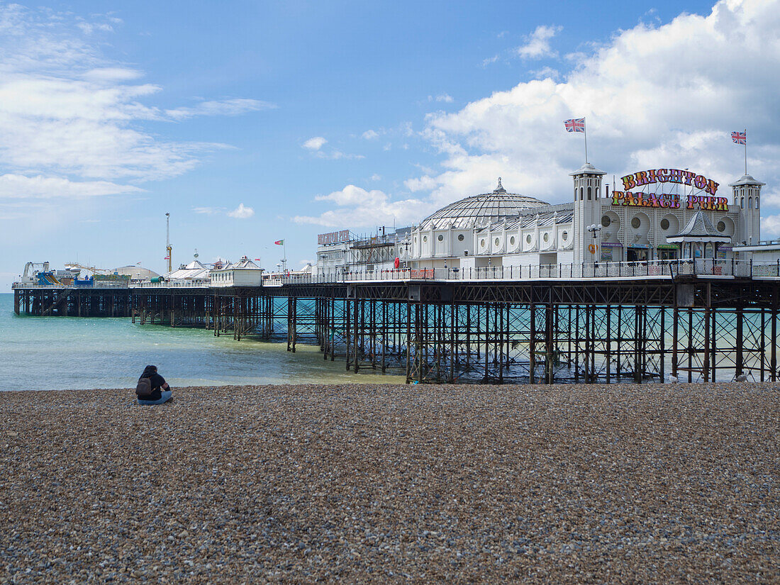 Blick auf den Brighton Palace Pier von Osten mit einer Frau, die am Kieselstrand sitzt,Brighton,East Sussex,England