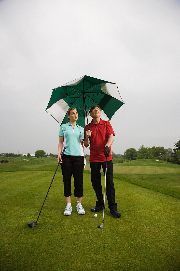 Vater und Tochter auf dem Golfplatz, einen großen Regenschirm haltend