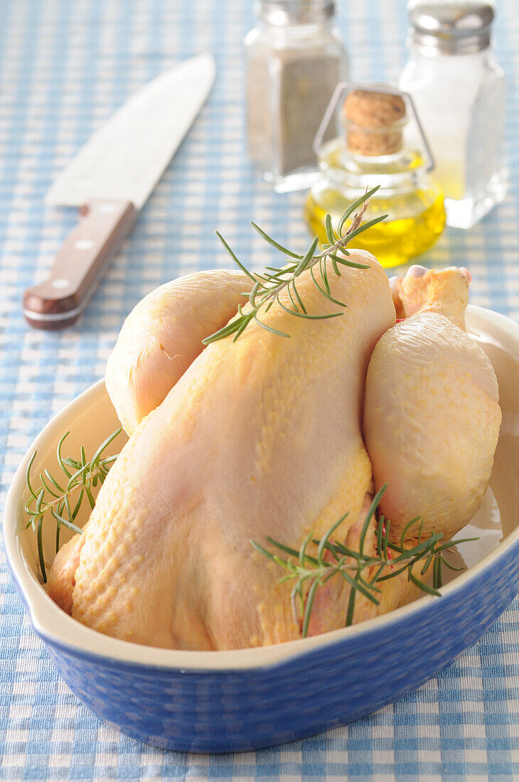 Ungekochtes ganzes Huhn mit Rosmarin in Bratpfanne auf blauem Gingham-Hintergrund, Studioaufnahme