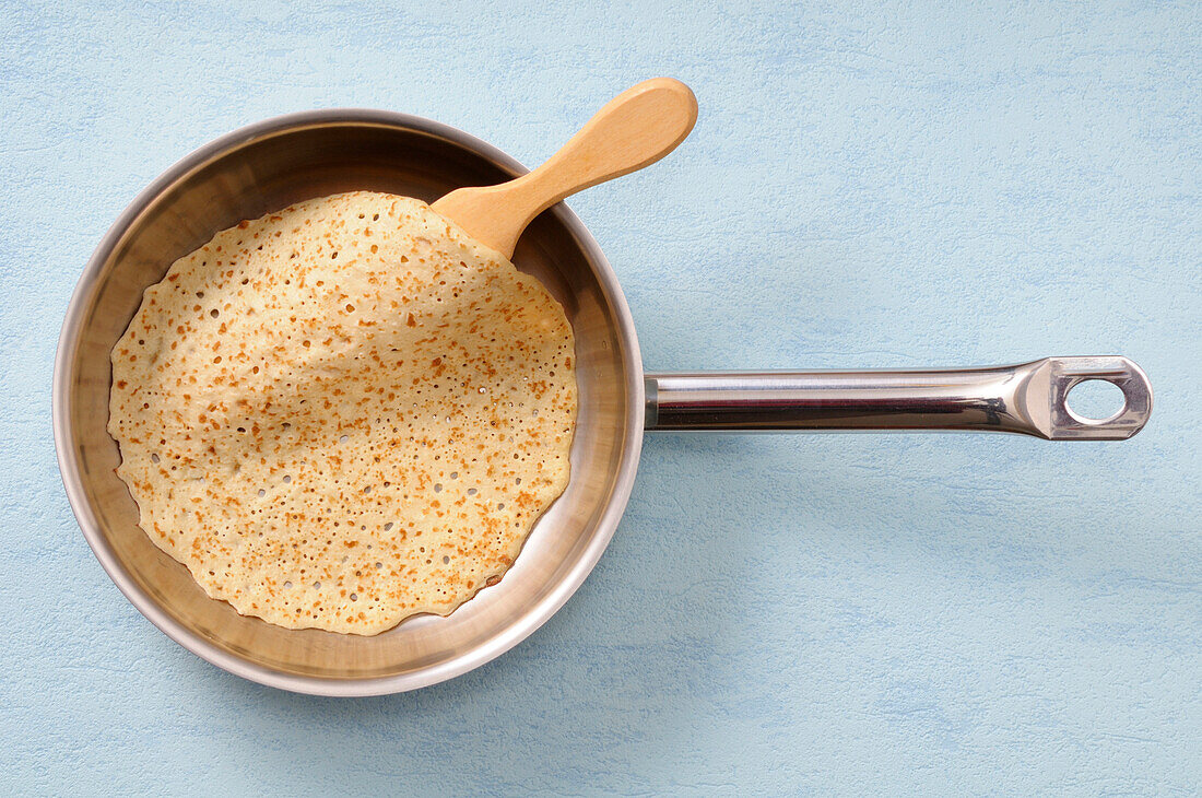 Draufsicht auf Pfannkuchen in Bratpfanne mit Spachtel auf blauem Hintergrund, Studioaufnahme