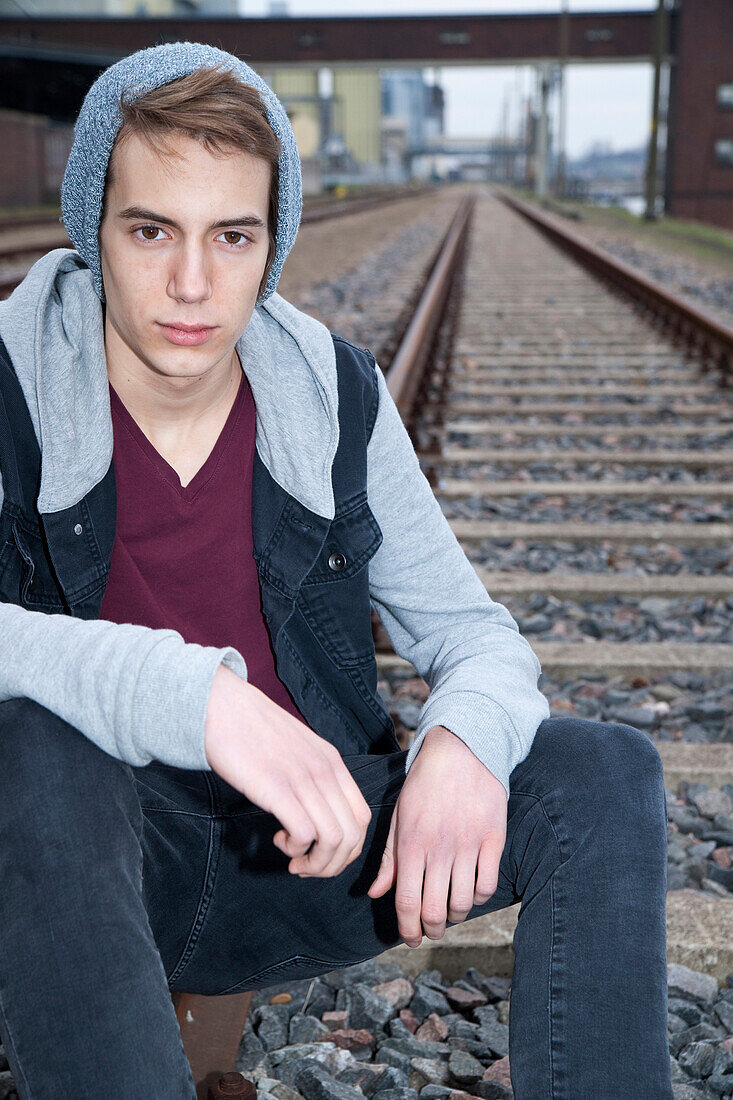 Porträt eines Teenagers auf Eisenbahnschienen sitzend, in die Kamera schauend, Deutschland