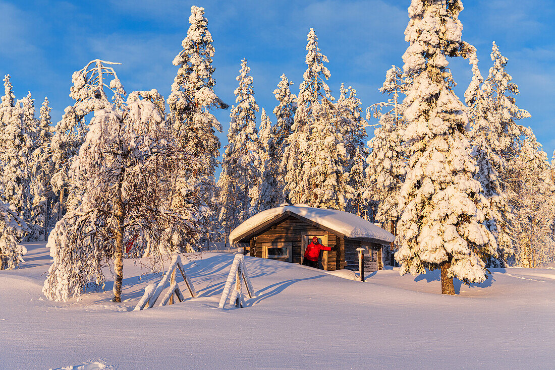 Tourist in der frühen Morgensonne steht vor einer isolierten Hütte im verschneiten Wald, Norrbotten, Schwedisch-Lappland, Schweden, Skandinavien, Europa