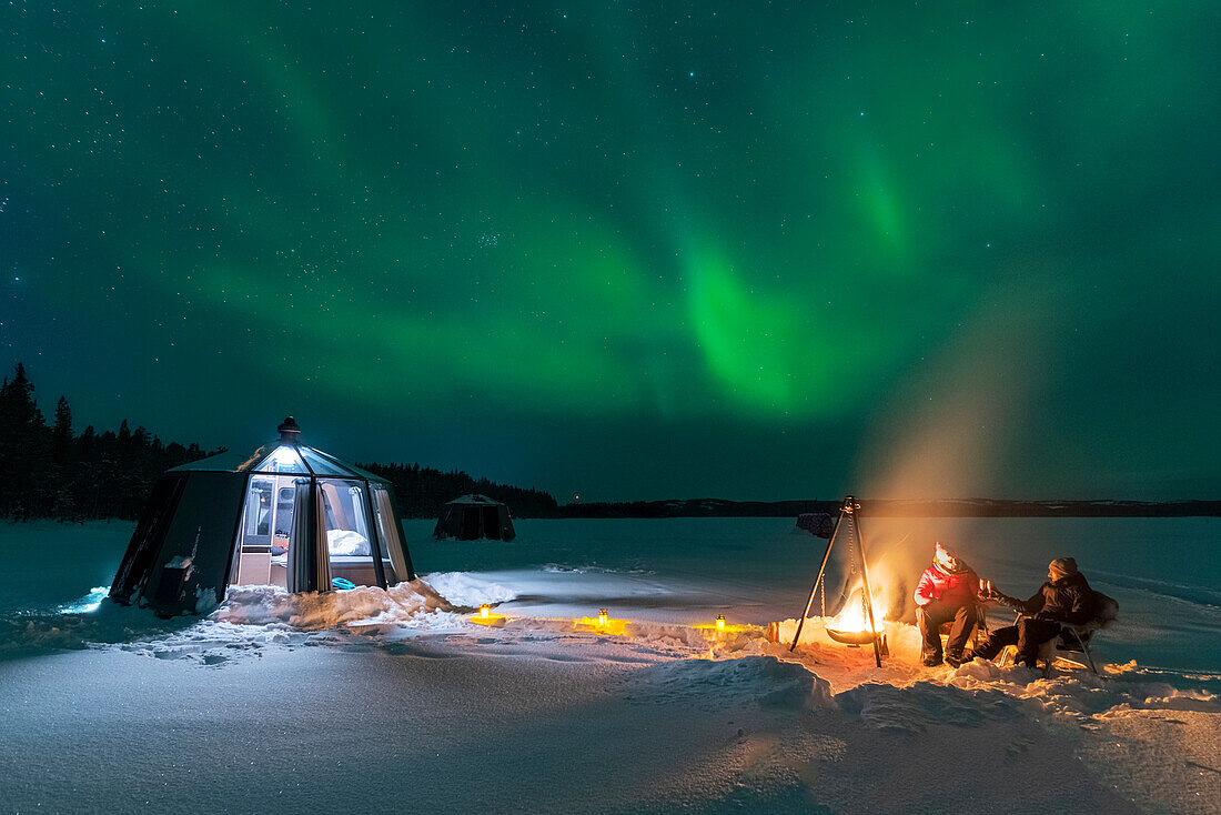 Night view of two people enjoyng dinner outside around a campfire close to the illuminated glass igloo with Northern Lights (Aurora Borealis) dancing in the sky,Jokkmokk,Norrbotten,Swedish Lapland,Sweden,Scandinavia,Europe