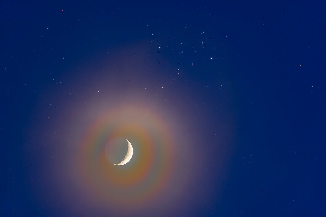The conjunction of the waxing 4-day-old crescent Moon below the Pleiades,and set in a slightly hazy sky on March 25,2023. The haze adds the colourful "lunar corona" halo around the bright crescent of the Moon from diffraction effects in the high icy clouds. Shot before the sky got dark,the remaining twilight adds the blue to the background sky. Earthshine is visible on the dark side of the Moon.