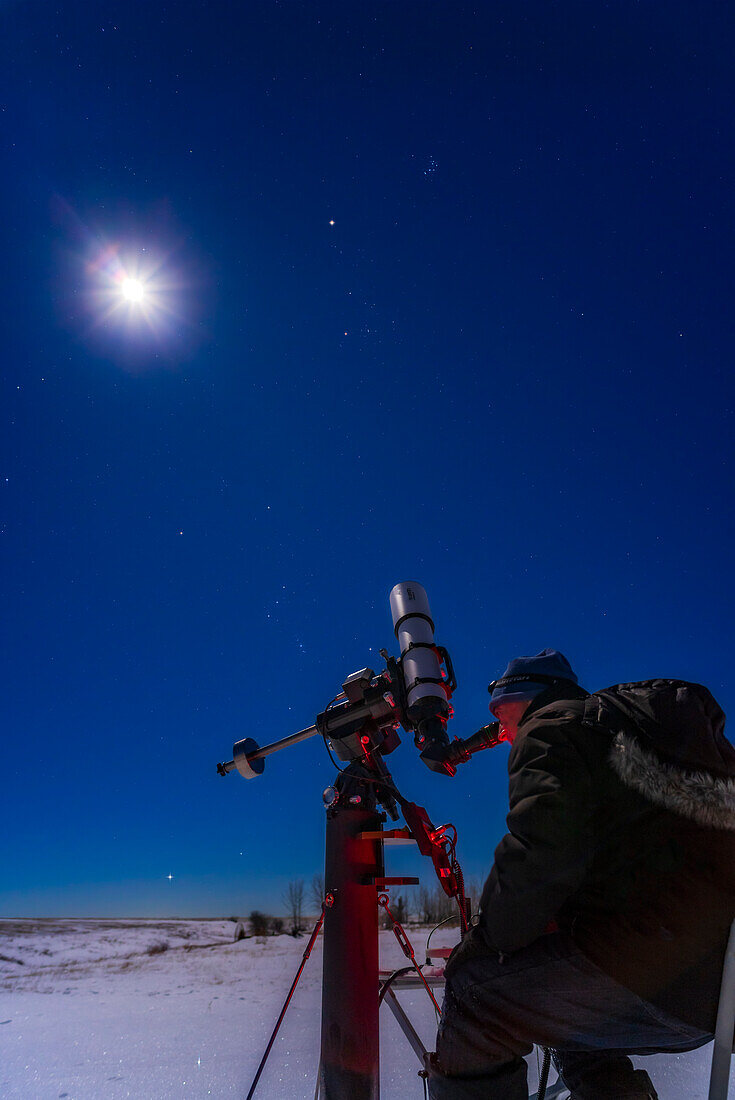 Ein Selfie von mir, wie ich Mars, den hellen rötlichen Stern oben, am 4. Januar 2023 von zu Hause aus beobachte, wobei Mars dann einen Monat nach der Opposition immer noch hell ist. Der zunehmende gibbous Mond ist das helle Objekt auf der linken Seite. Die Plejaden befinden sich oberhalb des Mars; die Hyaden und Aldebaran sind unterhalb des Mars. Orion und Sirius gehen im Hintergrund unten auf. Das Teleskop ist das Starfield Optics Gear115 auf der Astro-Physics Mach1-Montierung.