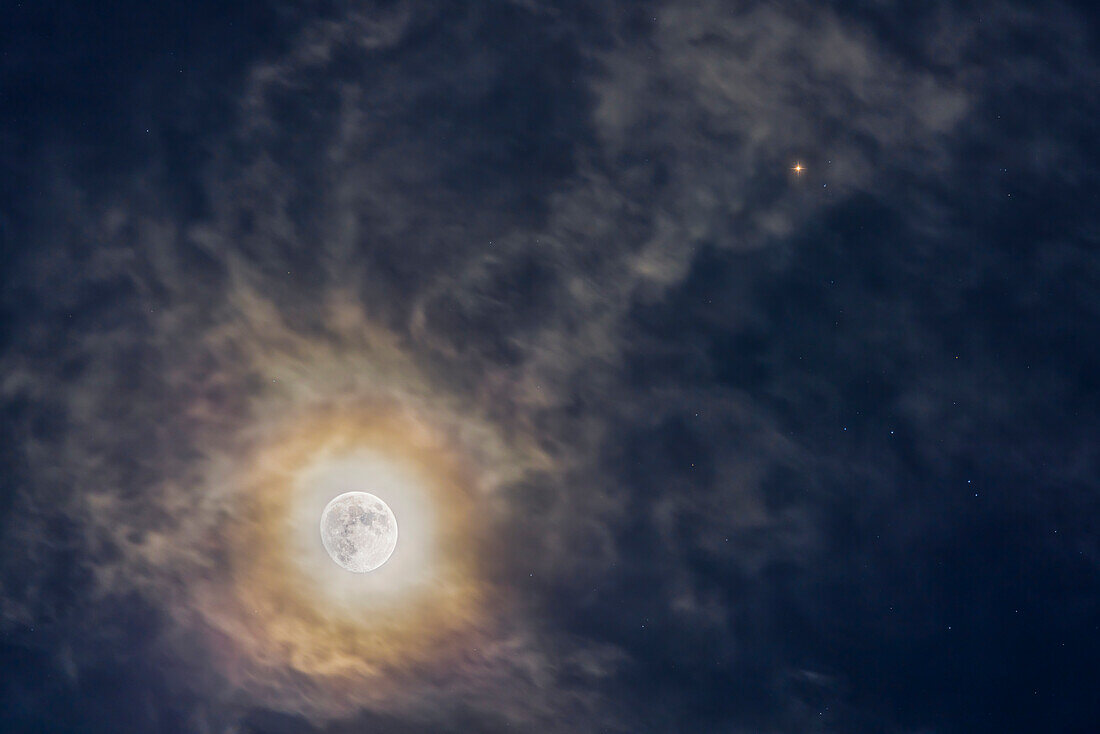 This is the waxing gibbous Moon (11.7 days old) near reddish Mars (at upper right),both set in a swirl of clouds,looking like they are in an interstellar nebula. Diffraction from ice crystals in the clouds adds the colourful corona around the Moon. This was the Moon-Mars conjunction of January 3,2023. Mars was then about a month past opposition.