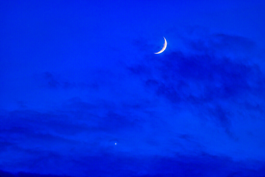 The conjunction of the waxing crescent Moon with Venus on June 21,2023,summer solstice evening,with the worlds in clouds in the deep blue twilight sky.