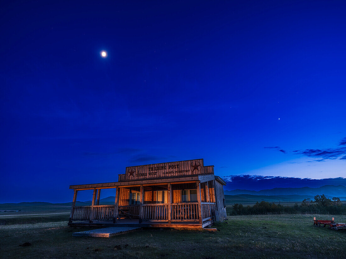 Der Viertelmond (in Löwe) und die Planeten Venus (am hellsten rechts) und Mars (schemenhaft und oben links von Venus) in der späten Abenddämmerung über den vorderen Bergketten der Rocky Mountains. Die Sterne Castor und Pollux in Zwillinge stehen über der Venus. Diese Aufnahme entstand am 27. Mai 2023 vom Grundstück Sierra Cabins West in der Nähe von Lundbreck, Alberta, während eines Workshops im Rahmen des Programms der Lightchasers Conference in Pincher Creek, Alberta. Die Beleuchtung erfolgt durch Mondlicht und einige Autoscheinwerfer.