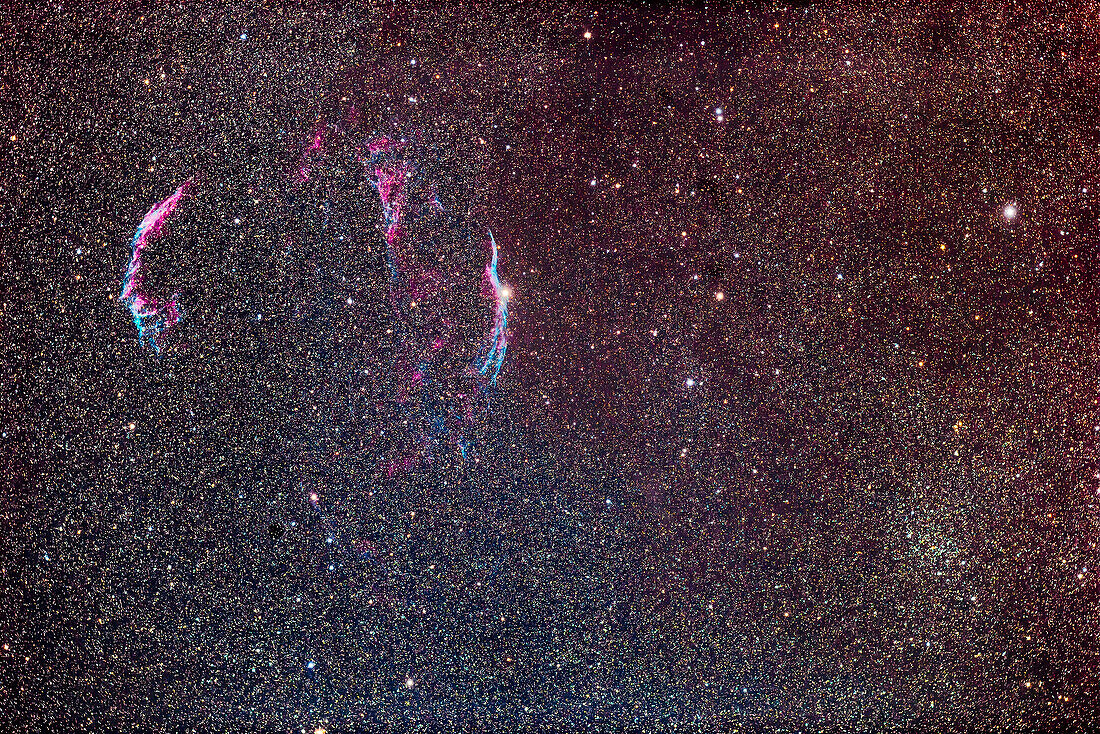 Die Bögen und Strähnen des Schleiernebels in Cygnus, eingerahmt von dem offenen Sternhaufen NGC 6940 in Vulpecula unten rechts. Das Gebiet ist durch einen scharfen Übergang zwischen klarem Sternenhimmel in der Millky Way und bräunlich-gelben Bereichen gekennzeichnet, die durch interstellaren Staub verdeckt sind und in denen weniger Sterne sichtbar sind. Die Farben des Nebels kommen gut zur Geltung, obwohl es sich um eine "normale" Kamera handelt, die keine Filter verwendet.