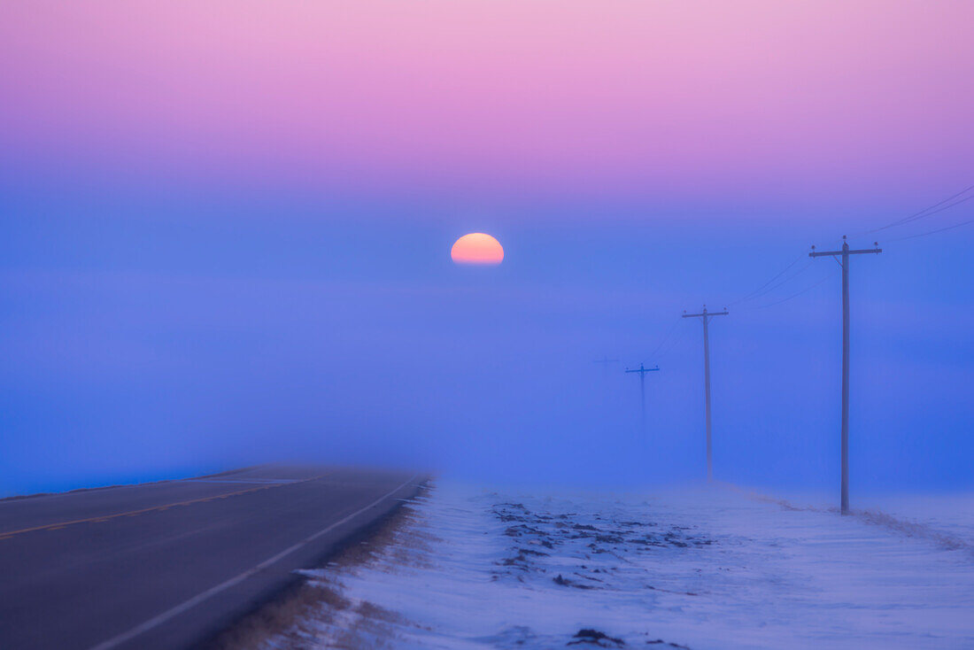 Die untergehende Sonne, die auf einer Landstraße in eine Nebelbank eintaucht, mit Blickrichtung Westen, am Abend des Frühlingsäquinoktiums, 20. März 2023. Die Sonne geht also genau im Westen unter. Der Nebel verdunkelt und rötet die Sonne, was die atmosphärische Absorption veranschaulicht. Dies war auf dem Highway 561 in Süd-Alberta.