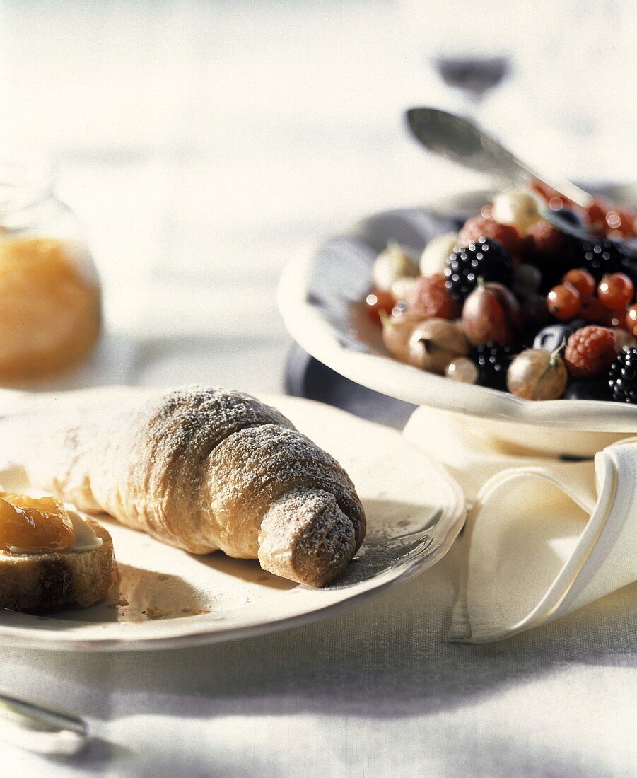Croissant auf Frühstücksteller, dahinter Schale mit Beeren
