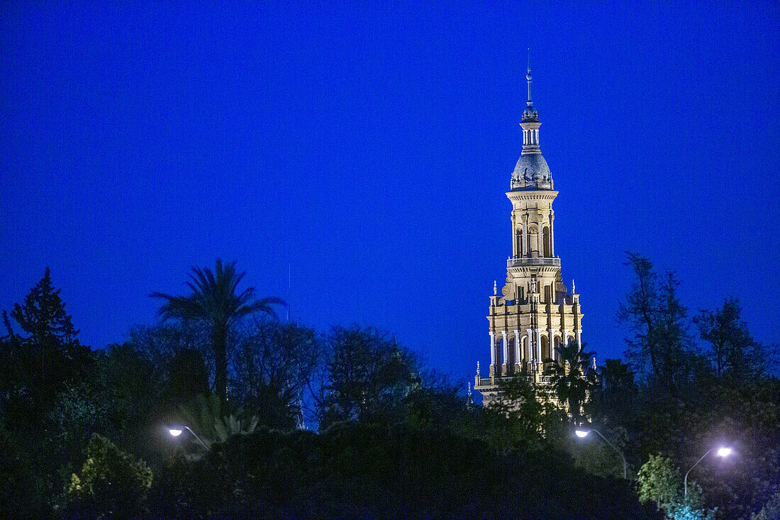 A stunning view of a tower of Plaza de Espana in Seville, Spain, illuminated against the night sky. The vibrant blue backdrop enhances the architectural beauty of this historic landmark.