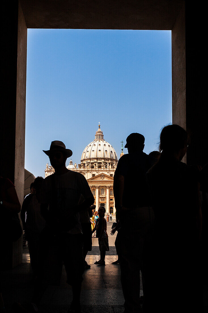 Tourists pass through an archway, revealing Saint Peter's Basilica under a clear sky in Vatican City, showcasing its grand architecture.