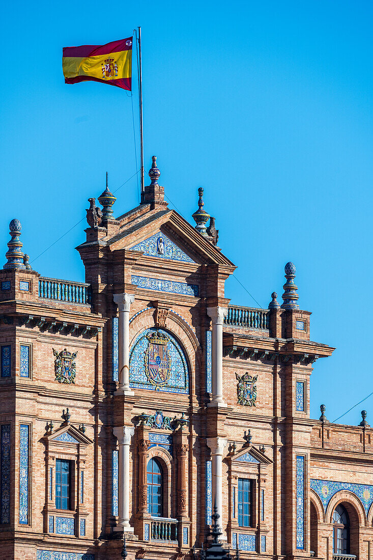 Nahaufnahme der komplizierten architektonischen Details der Plaza de España in Sevilla,Spanien,mit traditionellen spanischen Designelementen und einer leuchtenden Flagge vor einem klaren blauen Himmel.