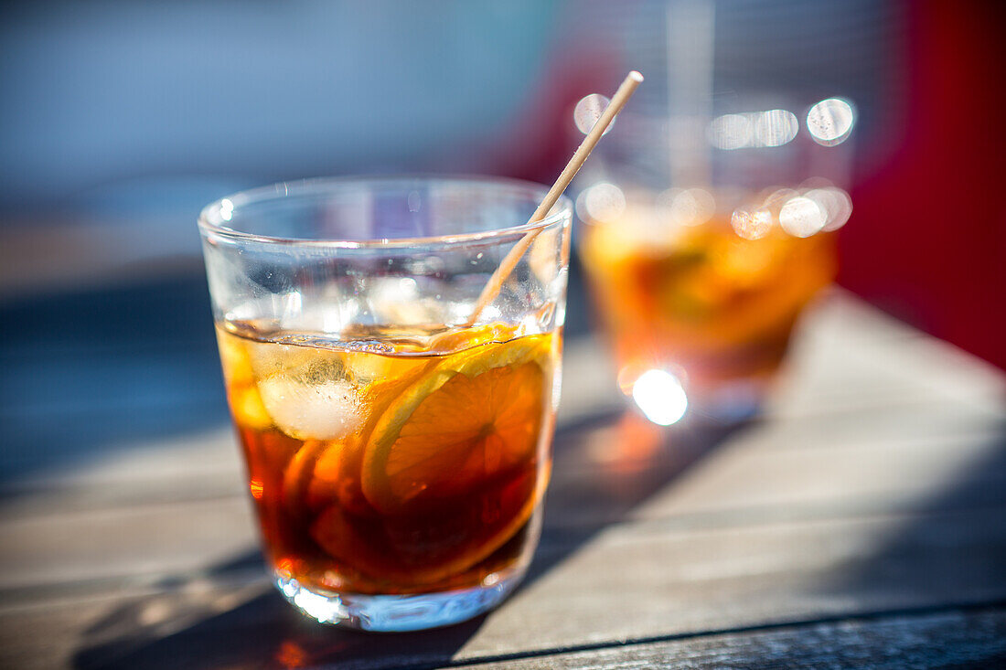 Eine Nahaufnahme von zwei Gläsern mit erfrischenden Wermut-Cocktails auf einem sonnenbeschienenen Terrassentisch,die die leuchtenden Farben und das einladende Ambiente hervorheben.