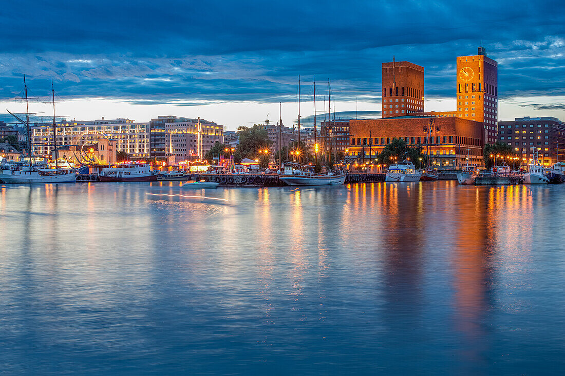 Stunning evening view of Oslo's City Hall and Aker Brygge from Akershus docks, with illuminated buildings reflecting on calm waters under a dramatic sky.