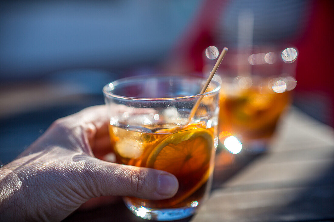 A man's hand holding a glass of vermouth with a slice of orange, capturing a refreshing and casual outdoor setting. Ideal for concepts of leisure, relaxation, and social gatherings.