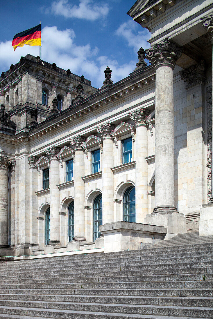 Visitors admire the impressive colonnade of the Reichstag building in Berlin under a bright blue sky adorned with a German flag.