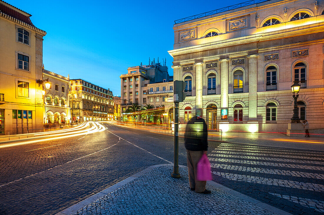 A long exposure capture of Dom Joao da Camara Square in Lisbon, Portugal, featuring the illuminated National Theatre in the foreground during evening hours.