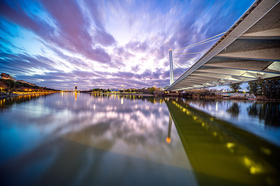 Beautiful dusk scene capturing the illuminated Alamillo Bridge over Rio Guadalquivir in Sevilla, Spain. The reflection on the water adds tranquility to the vibrant cityscape.