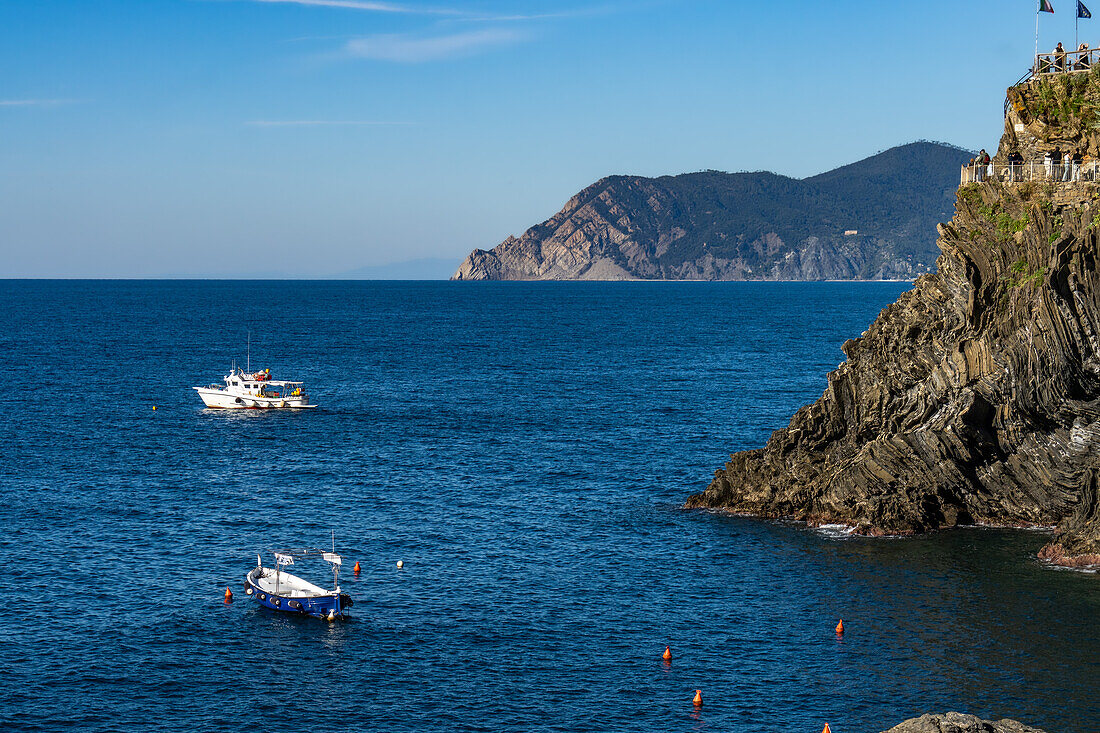 Fischerboote vor der zerklüfteten Küste der Stadt Manarola in den Cinque Terre, Italien. Touristen befinden sich auf einem Aussichtspunkt rechts.