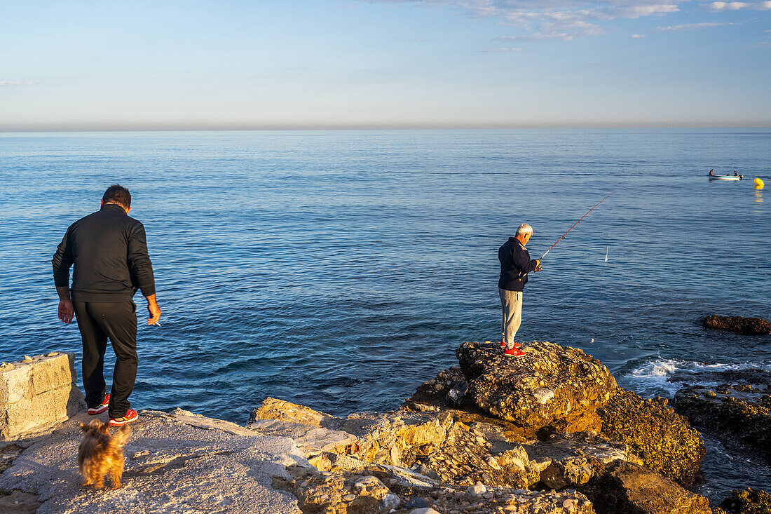 Two men enjoying fishing on a rocky shoreline overlooking the calm Mediterranean sea.