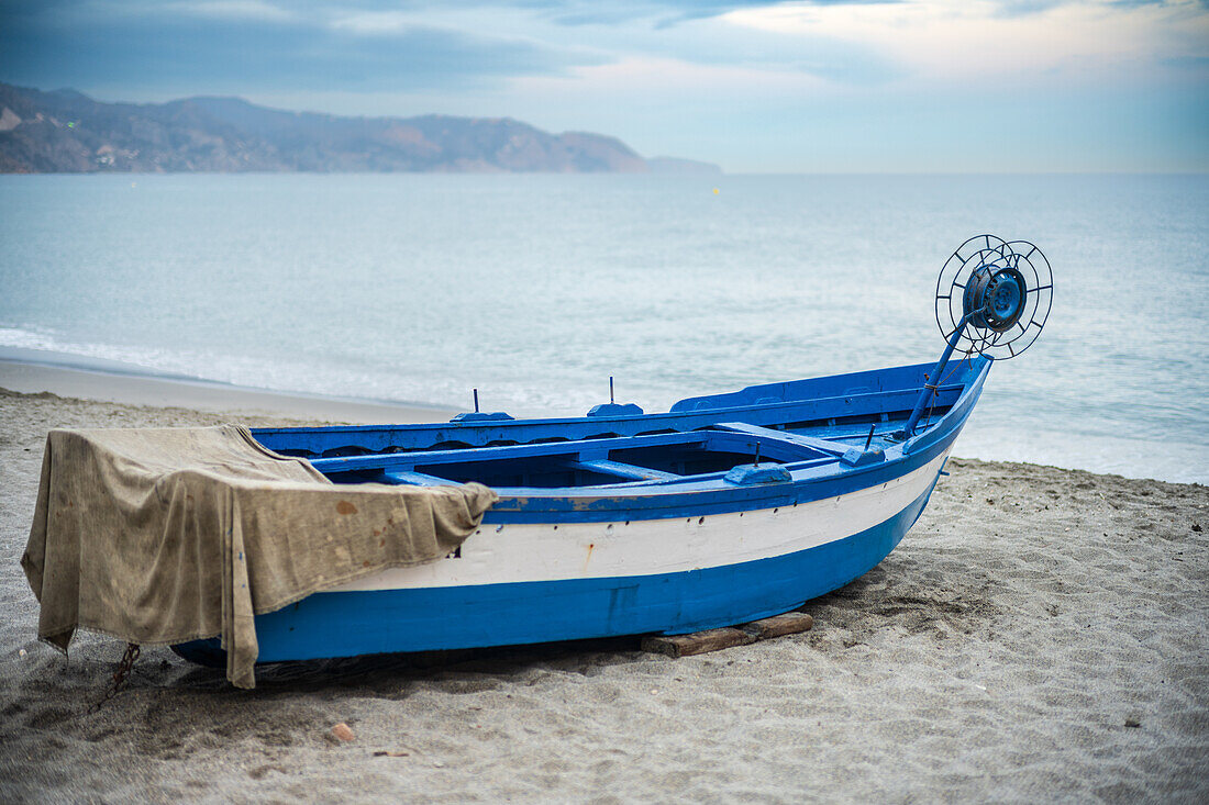 A blue fishing boat rests on a sandy shore under overcast skies near the coastline.