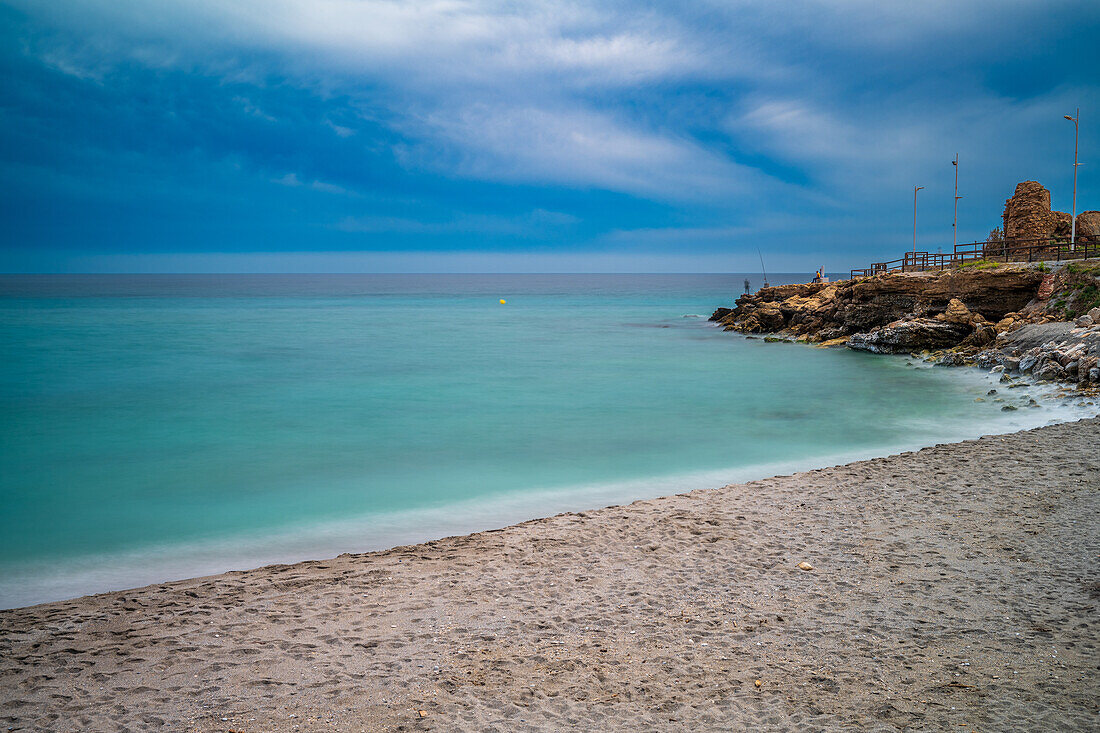 Long exposure photo capturing Torrecilla Beach with a tranquil sea and rocky shore.