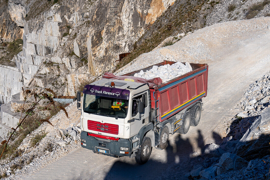 Ein Lastwagen, der Marmor transportiert, während der Besichtigung eines Steinbruchs in Fantiscritti. Carrara, Italien. Die Schatten der Teilnehmer sind auf der Straße zu sehen.