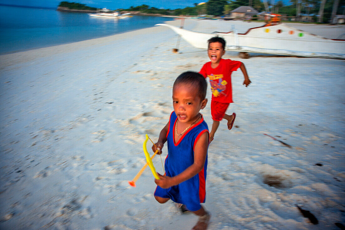 Einheimische Kinder spielen mit Pfeil und Bogen am weißen Sandstrand von Langub Beach auf der Insel Malapascua, Cebu, Philippinen.