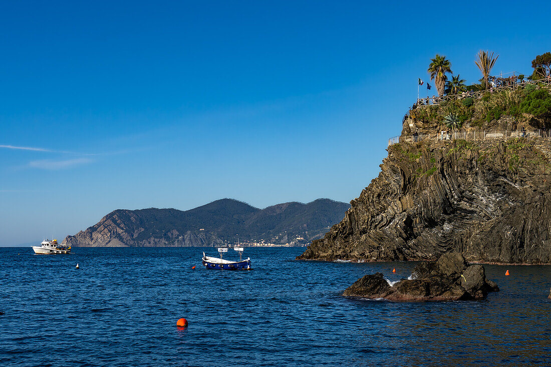 Fischerboote vor der zerklüfteten Küste der Stadt Manarola in den Cinque Terre, Italien. Touristen befinden sich auf einem Aussichtspunkt rechts.