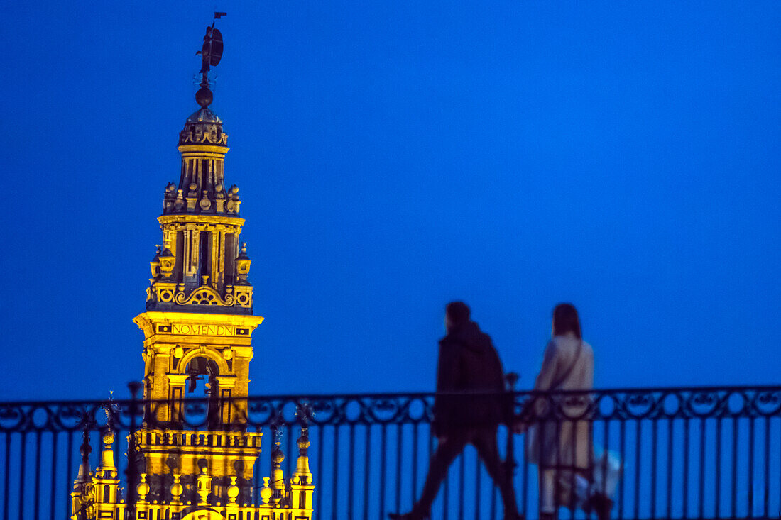 People walking on a bridge with Giralda Tower background in Seville during twilight.