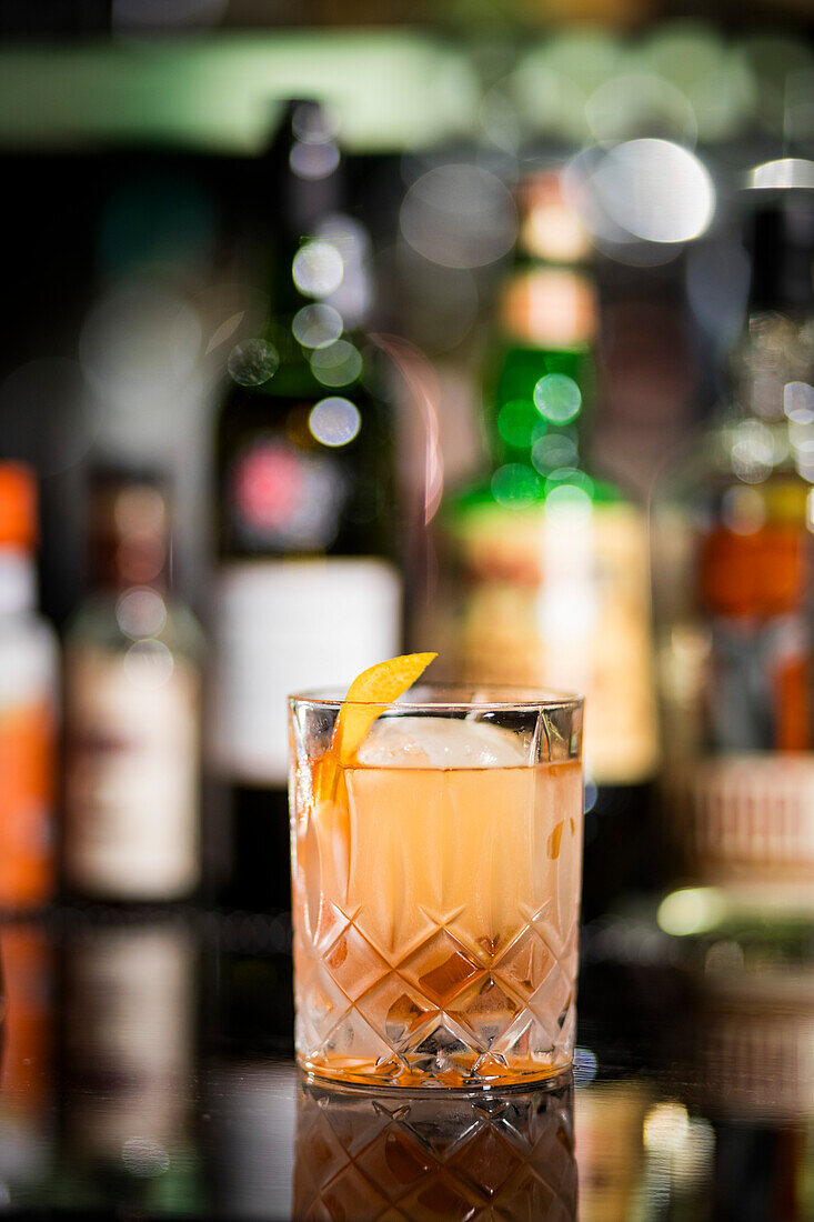 Alcohol cocktail with ice and lemon twist, liquor bottles in background