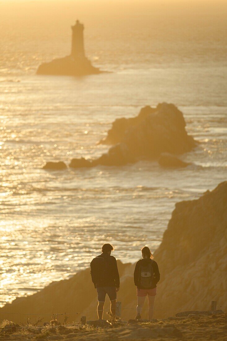 Frankreich, Finistere, Plogoff, Wanderer bei Sonnenuntergang an der Pointe du Raz, im Hintergrund der Leuchtturm der Vieille