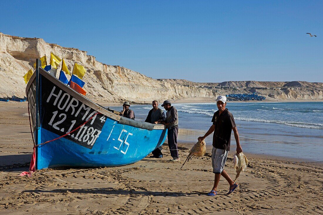 Marokko, Westsahara, Dakhla, Fischer entladen Fisch in der Nähe ihres blauen Bootes am Strand von Araiche, der von einer Klippe begrenzt wird