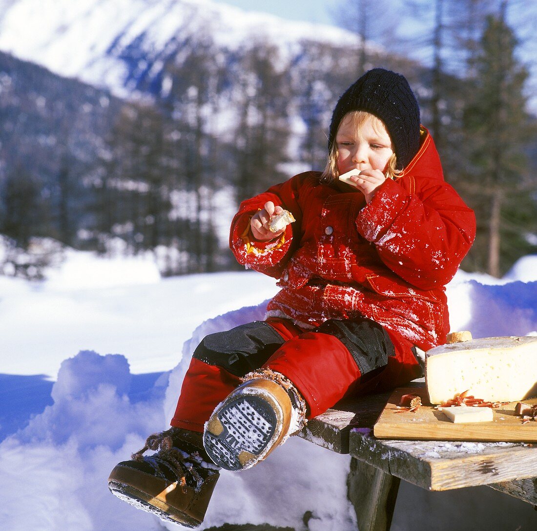 Child in winter clothes at table in snow eating bread & cheese