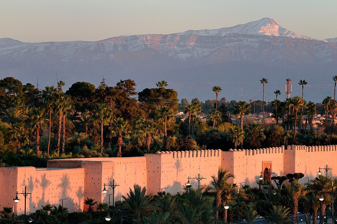 Marokko, Hoher Atlas, Marrakesch, die von der UNESCO zum Weltkulturerbe erklärte Kaiserstadt Medina, die Stadtmauern und der Oukaïmeden-Gipfel im schneebedeckten Atlas im Hintergrund bei Sonnenuntergang