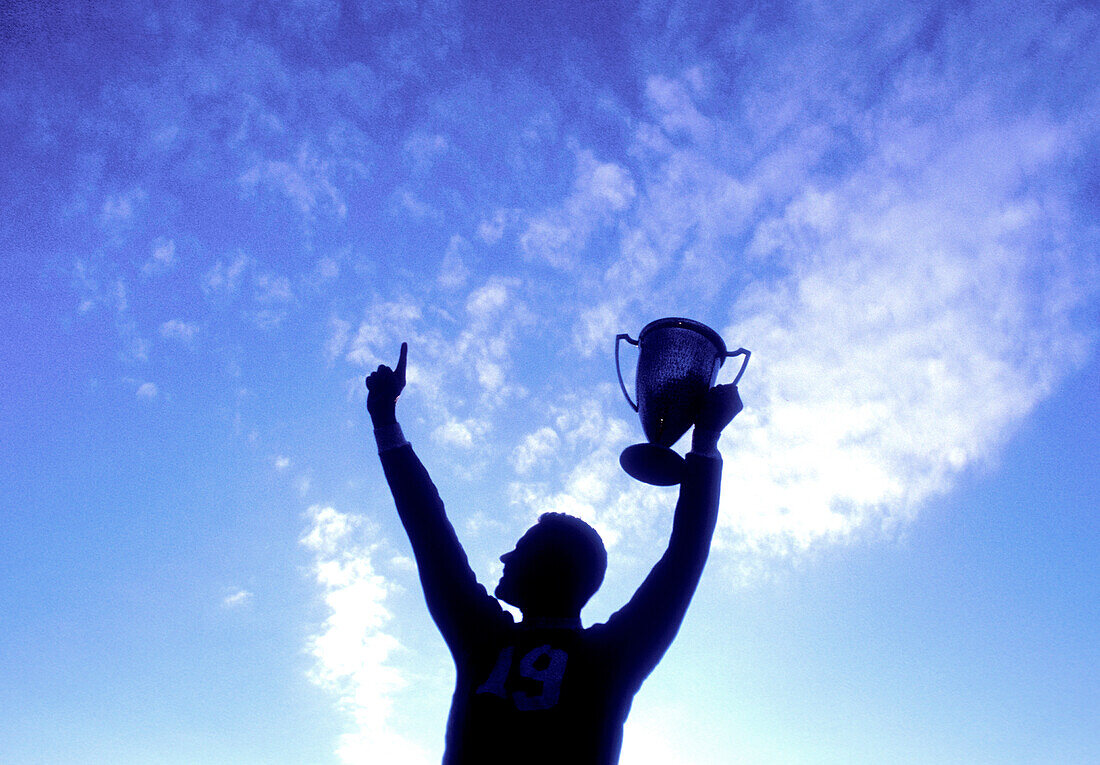 Silhouette of man holding up trophy against sky