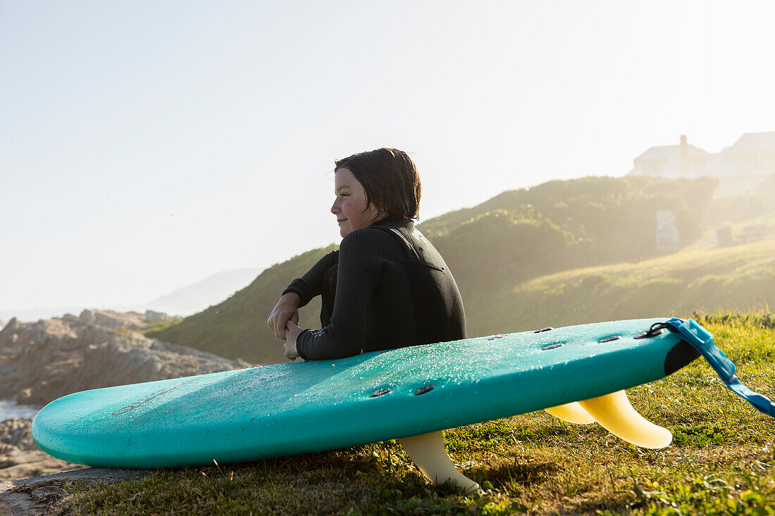 Junge (10-11) mit Surfbrett sitzend am Kammabaai Beach