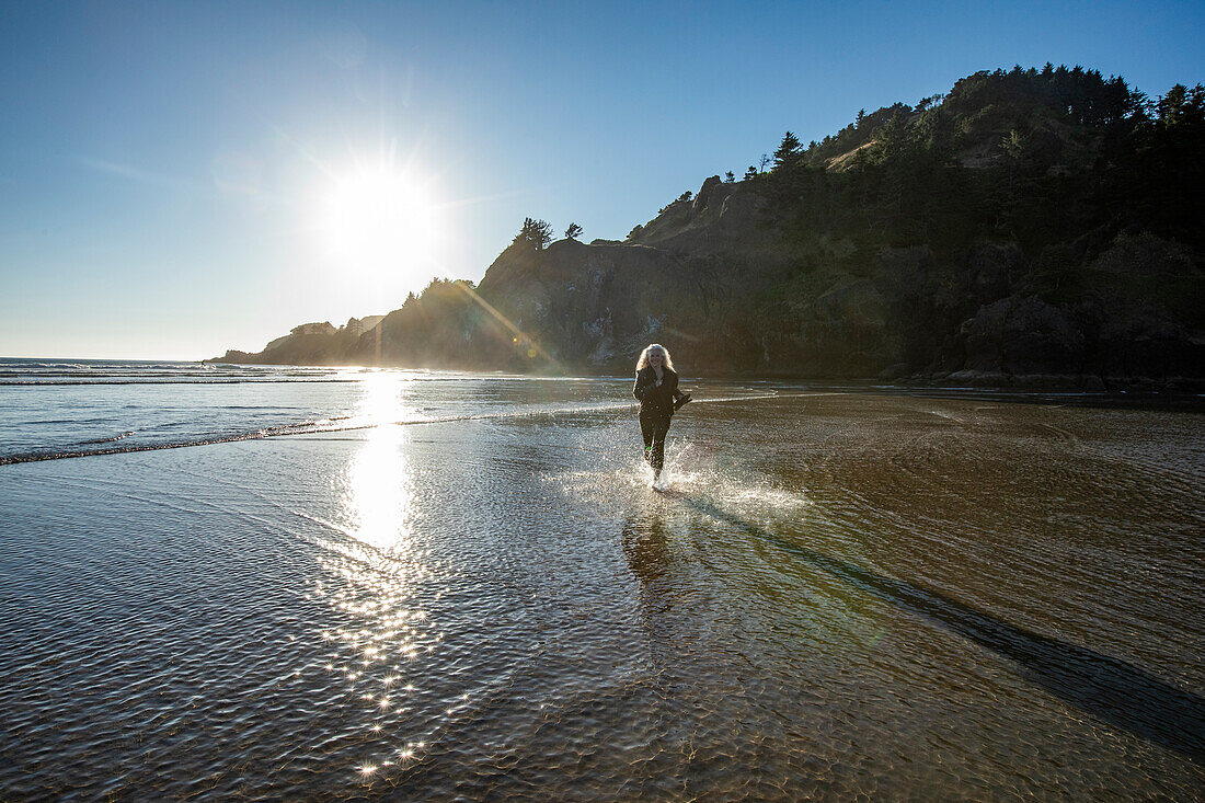 USA, Oregon, Newport, Frau läuft am Sandstrand und spritzt Wasser