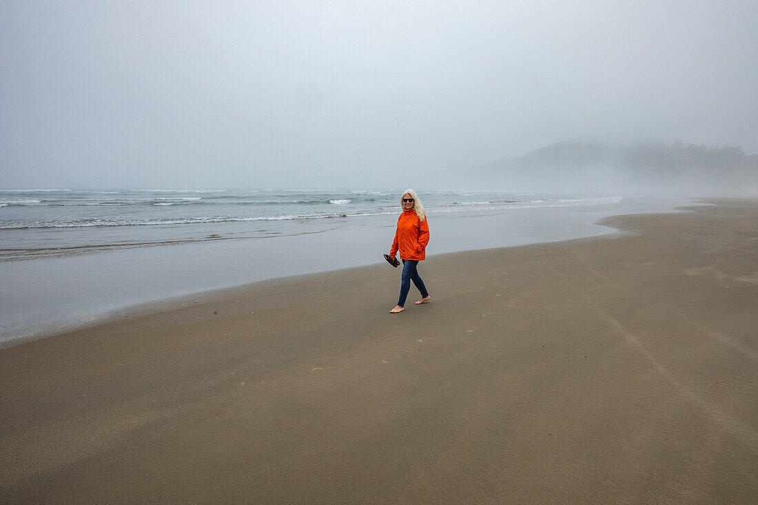 Frau in orangefarbener Jacke geht am nebligen Strand entlang