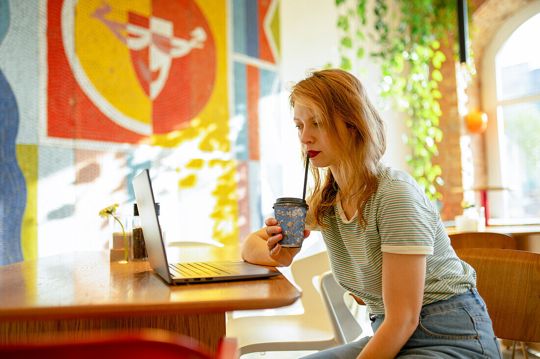 Seitenansicht einer Frau, die an einem Laptop arbeitet, während sie Kaffee im Café trinkt