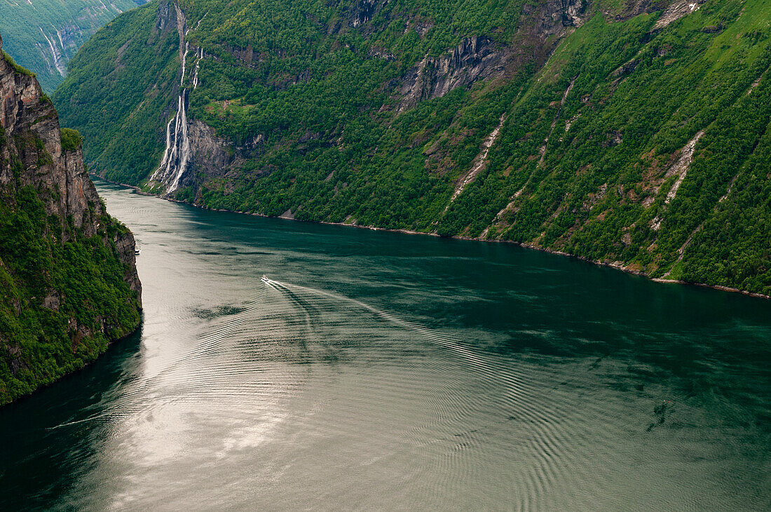 Das Kielwasser eines Bootes kräuselt den Geirangerfjord, der sich durch waldbedeckte steile Klippen und Berge schlängelt. In der Ferne sind die Sieben-Schwestern-Wasserfälle zu sehen. Geirangerfjord, Norwegen.