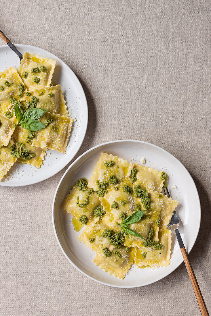 Draufsicht auf appetitlich gekochte Ravioli mit grüner Soße und Kräutern auf weißen Tellern mit Gabeln auf dem Tisch