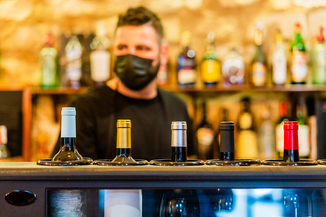 Unscharfer Barmann mit steriler Maske steht am Bartresen, während er in einem Restaurant während des Coronavirus arbeitet