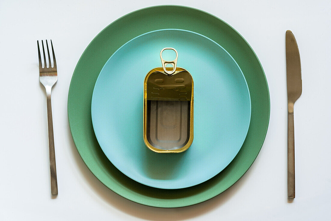 Draufsicht auf eine geöffnete leere Metalldose mit aufklappbarem Deckel auf einem blauen Keramikteller, der auf einem weißen Tisch mit silbernen Gabeln und Messern serviert wird
