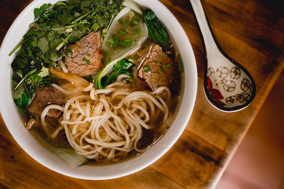 Von oben große weiße Keramikschüssel mit Rindfleisch-Ramen-Suppe und buntem Löffel auf Holztisch platziert